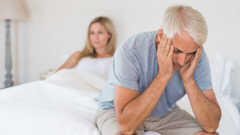 A man sits on the edge of a bed with his hands cradling his face. A woman sits under the covers in the background.