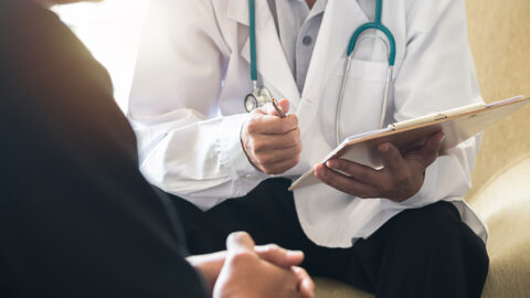 A doctor holding a clipboard and pen talks to a male patient.