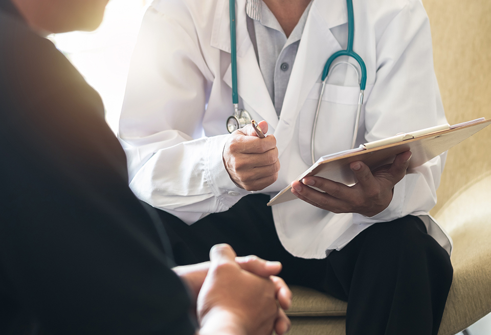 A doctor holding a pen and clipboard talks to a male patient.