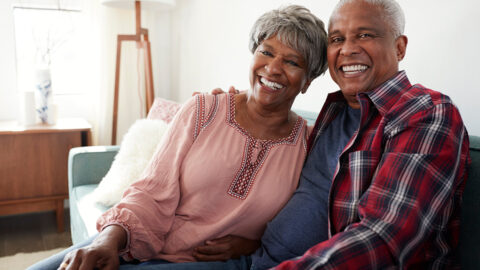 A woman and a man sit on a couch, smiling toward the camera. The woman leans her head against the man, who has an arm around her shoulders.