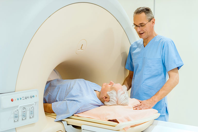 A medical technician stands next to a patient who is laying on a table receiving a medical scan in an MRI machine