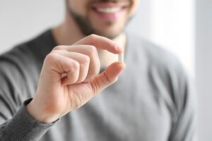 Closeup on the hand of a man holding a small white pill between his thumb and index finger.