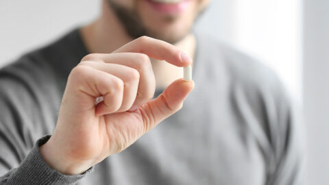 Closeup on the hand of a man holding a small white pill between his thumb and index finger.